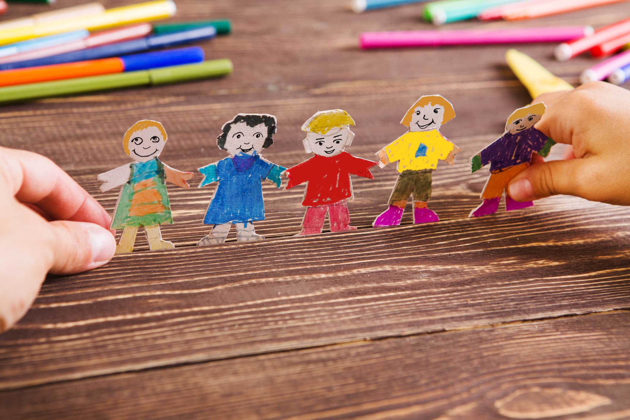 Children's hands holding a chain of hand-drawn paper dolls on a wooden table scattered with markers